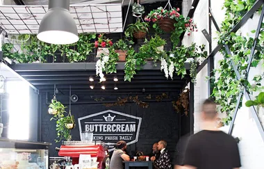 Indoor cafe scene with hanging plants, people sitting, and a sign reading "Buttercream Baking Fresh Daily."