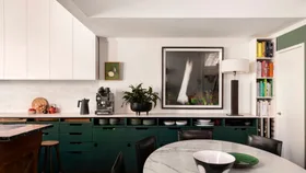 A kitchen with white countertops, green cabinets underneath the counter, white cabinets above the counter and a white tiled splashback. Coffee machine, plants and a lamp are on the countertop, while art is on the wall. A round marble table is positioned in front.