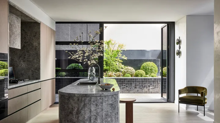 A kitchen that leads out on to an outdoor space. A large island is centred in the kitchen, with grey marble on the top and sides. This marble is also on the splashback, rangehood and benchtop, surrounded by Eveneer joinery.