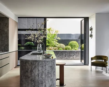 A kitchen that leads out on to an outdoor space. A large island is centred in the kitchen, with grey marble on the top and sides. This marble is also on the splashback, rangehood and benchtop, surrounded by Eveneer joinery.