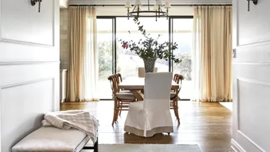 Dining room with wood table, chairs, flower centerpiece, large windows, light curtains, and a bench with a blanket.