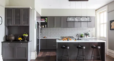 Modern kitchen with grey cabinets, patterned backsplash, white counters, and industrial pendant lights over an island.