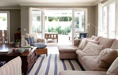 Bright living room with beige couches, striped rug, wooden coffee table, and large windows opening to a green garden.