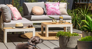 Outdoor patio with gray seating, pink pillows, a straw hat, sunglasses on a table, and a dog in foreground.