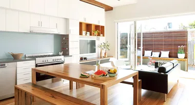 Bright modern kitchen-living space with wooden table, fruit bowl, black sofa, and glass doors opening to patio.