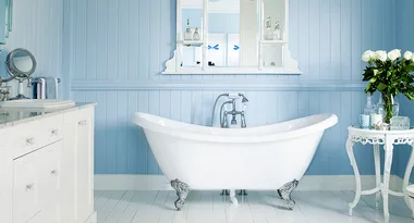 Elegant bathroom with a white clawfoot tub, blue panel walls, mirror, vanity, and table with flowers.