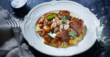 A plate of lamb tagine with chickpeas, almonds, and cilantro served over couscous, next to a fork and glass.