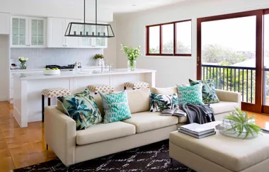 Modern living room with beige sofa, colorful pillows, open kitchen, and a large window view.