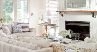 Bright living room with white sofas, blue cushions, a mirror, fireplace, and a wooden table holding books and an orchid.