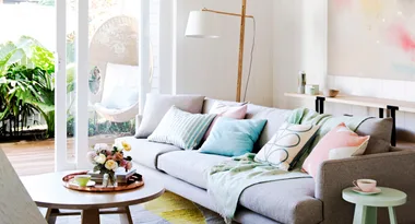 Cozy living room with a gray sofa, pastel pillows, and a coffee table with flowers, opening to a lush garden view.