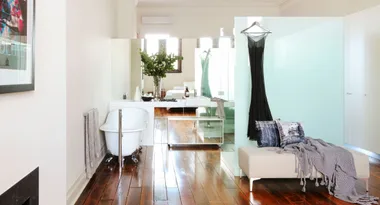 Luxurious bathroom with bathtub, elegant black dress hanging, modern sink, vase with greenery, and cozy seating area.