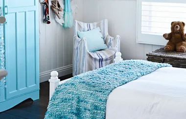 Cozy bedroom with blue accents, a teddy bear on a wicker chest, and a striped chair near a wardrobe.