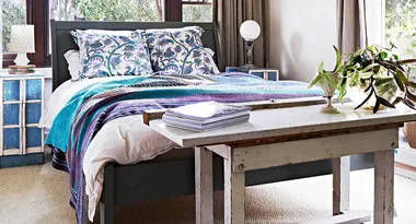 Cozy bedroom with a patterned bedspread, pillows, bedside tables, and a rustic wooden bench with plants and books.