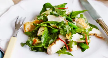 Grilled fish salad with fresh greens, herbs, and sliced red chili on a white plate, fork and knife on the side.