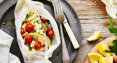 Fish baked in parchment with lemon, cherry tomatoes, and herbs, served on a plate with a fork and knife.