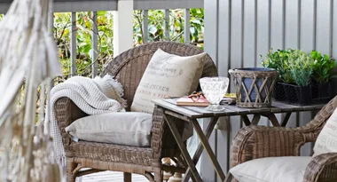 Wicker chair with cushions and blanket on a porch, beside a wooden table with plants and decor items.