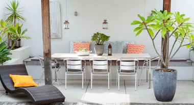 Outdoor patio with a dining table, metal chairs, accent pillows, hanging lanterns, and large potted plants.