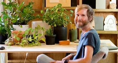 A person with a beard sits at a desk surrounded by potted plants, smiling and wearing a blue shirt.