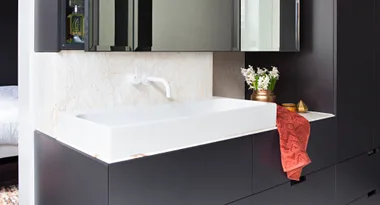 Modern bathroom with black cabinets, white rectangular sink, red towel, brass vase with flowers, and gold decor.