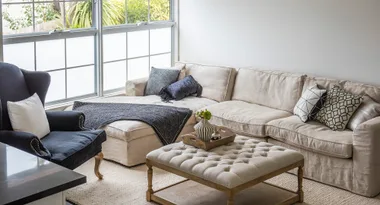 Living room with beige sectional sofa, patterned cushions, navy chair, tufted ottoman, and large windows.