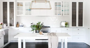 White kitchen with glass cabinets, a central island with plants, and modern lighting fixtures above.