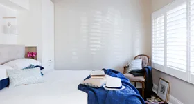 Cozy bedroom with white bed, blue blanket, book, hat, wooden chair, and sunlight through shuttered windows.