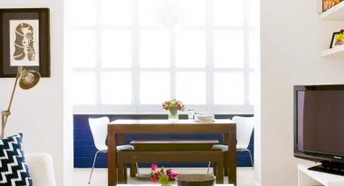 Dining area with dark wooden table, chairs, flowers, and bright windows; art, lamp, and TV visible in room.