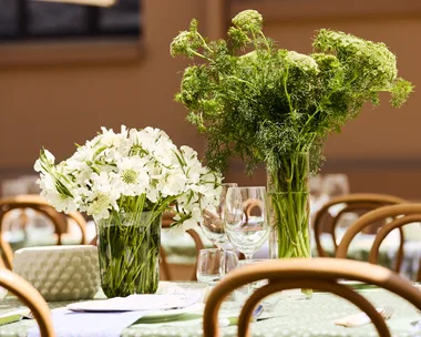 Elegant table setting with vases of white and green flowers, surrounded by wooden chairs and glassware.