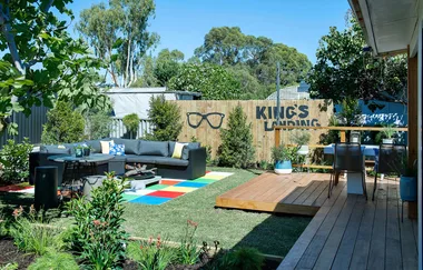 Backyard with colorful patio, L-shaped sofa, "King's Landing" on fence, dining set on wooden deck, and lush greenery.