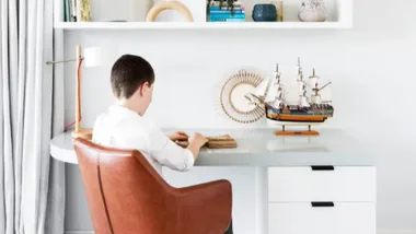 A child sits at a desk with a model ship, books, and decorative items on shelves in a bright room.