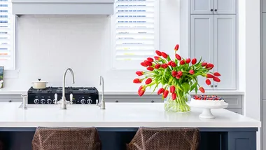 Modern kitchen with white countertops, red tulips in a vase, fruit-topped cake, and wicker chairs.