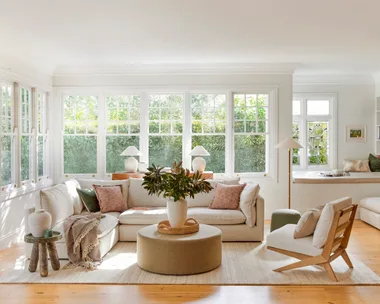 A living room with light timber floorboards, white walls and large windows that look out on to greenery. A cream modular sofa is positioned in the middle, styled with throws and cushions. A round ottoman is positioned in the middle, creating a coffee table that has a cream vase on top, displaying greenery.