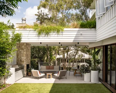 A white exterior of a home. A stone clad fireplace has a chimney that reaches to the white terrace that stretches above the alfresco barbecue kitchen and dining area. There's a strong indoor outdoor connection with alfresco dining and alfresco lounge furniture with a barbecue.