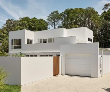 Modern white two-story house with large windows and a garage, surrounded by trees and a small fence.