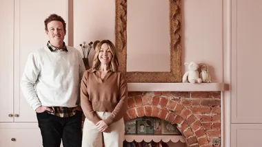 Two people stand in front of a pink wall with a mirror and brick fireplace, decorated with toys and books.