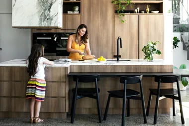 Woman cutting bread and child watching in modern kitchen with wooden cabinets and marble countertops.