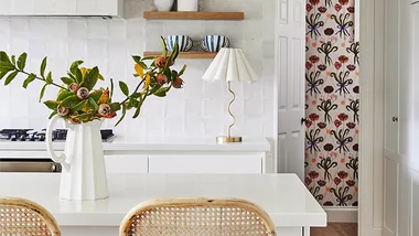 Kitchen with white island, wicker chairs, floral vase, patterned wallpaper, and a lamp on the counter.