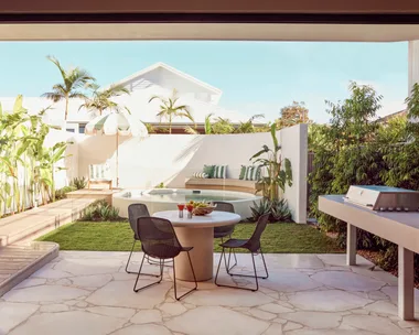 An alfresco dining area with a barbecue with a curved white wall around a circular plunge pool in the background. Green and white striped cushions decorate the bench around the pool and stone is used for the flooring of the outdoor dining area.