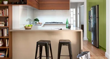 Modern kitchen with wooden cabinets, green accent wall, metal stools, and bookshelves. Bright, airy space with a countertop.