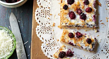 Blackberry coconut bars on a wooden board with a doily, beside grated coconut and a knife.