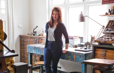 A woman stands by a wooden desk in a well-lit workshop with art supplies and books.