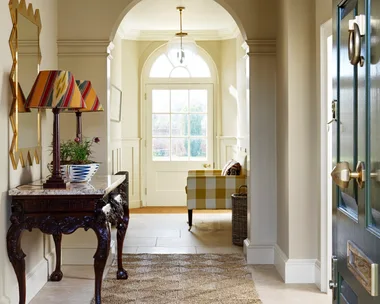 A hallway entrance to a home, with tiled flooring and a rug placed atop. This liminal space has a console table with two lamps on top and a gold-framed mirror on the wall.