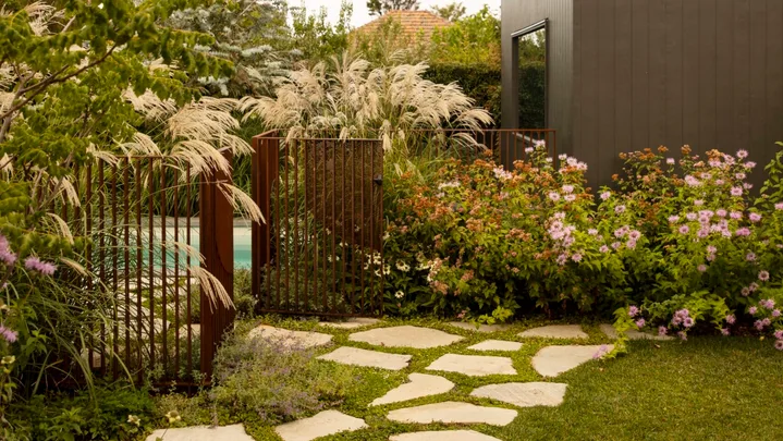 Modern garden with stone paver path, lush plants, and a dark grey building under a bright, cloudy sky. The result of a garden makeover.