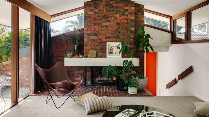 Mid-century style living room with red brick fireplace, brown chair, plants, and a large window with trees outside. Grey carpet is in front of the brick with a round coffee table and scattered cushions around.