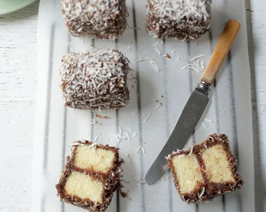Lamingtons coated with chocolate and coconut on parchment paper, accompanied by a knife and a cup of tea.