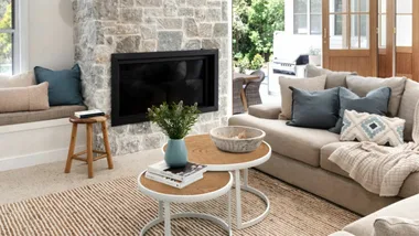 Cozy living room with a stone fireplace, beige sofa, blue cushions, and a round coffee table with decor.