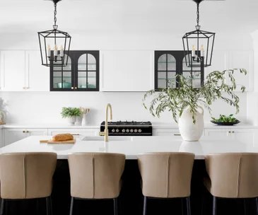 Modern kitchen with white cabinets, black stove, large island with beige chairs, two pendant lights, and a plant on the counter.