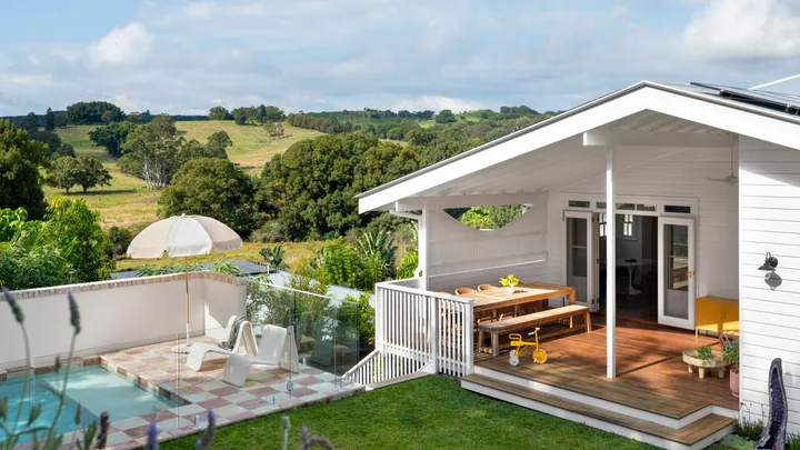 White house with wooden deck, outdoor dining table, small pool, umbrella, chairs, and scenic countryside view.