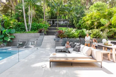 A modern outdoor patio with lush greenery, seating area, and a pool. Three people are enjoying a sunny day.