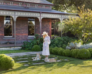 Woman in floral dress and hat standing in a garden with two corgis, brick house in the background. The garden was designed by Paul Bangay.